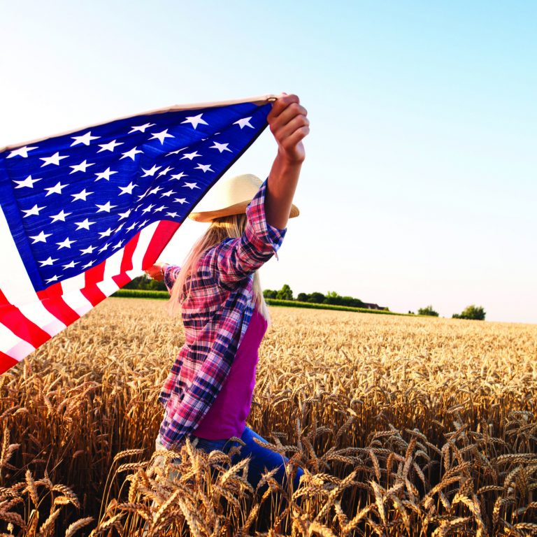 Concept of freedom and liberty. Attractive blonde female walking through wheat field and holding USA waving flag. Powerful economy and agriculture of the United States of America. Proud on her country