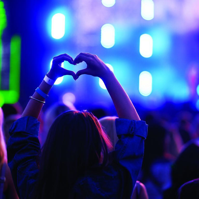 Person forms heart shape with hands at lively outdoor music festival, blurred stage lights, crowd enjoys event. Love symbol gesture, fans celebrate artist, youth culture, happy summer vibe in concert.