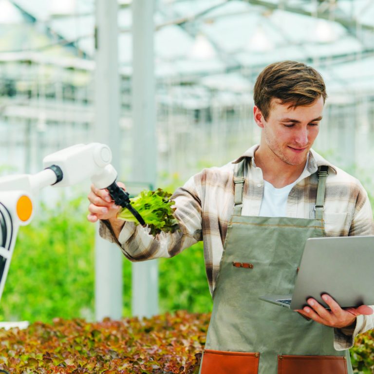 Male farmer using laptop in greenhouse to check the operation of robotic arm with AI system to control in farm care. Smart agriculture technology concept. Wireless robot for agriculture in smart farm.